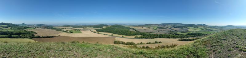 Panoramatický pohled z vrchu kopce Číčov, je to kamenitý kopec porostlý řídkou vegetací, v dálce jsou vidět pole, lesy, kopce, některé charakteristicky špičaté pro České Středohoří
