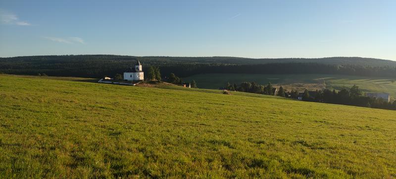 Svažitá zelená louka ze které vystupuje kostel se hřbitovem osvětlený odpoledním sluncem