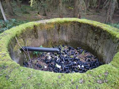 Betonová skruž v lese, několik metrů vysoká a dva metry v průměru, do které je zavedena plastová trubka zřejmě na napouštění vodou. Skruž je plná kousků rozřezané černé vrubované trubky.